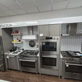 Italian-inspired Bertazzoni kitchen appliance wall featuring a collection of professional-grade ranges, cooktops, built-in ovens, and a panel-ready refrigerator, presented with a stylish geometric backsplash and modern shelving.
