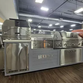 Premium Lynx outdoor kitchen setup featuring a full stainless-steel grilling station, built-in refrigerator, pizza oven, and side burners on display inside Art Handler’s Appliance showroom.