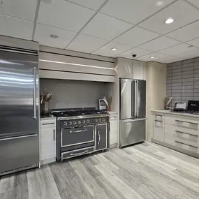 A modern luxury kitchen display featuring premium Viking appliances, including a side-by-side refrigerator, French door refrigerator, and a professional-grade dual oven gas range, set in a contemporary showroom kitchen layout.