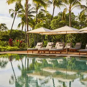 Pool surrounded by palm trees and lounge chairs near the ocean.