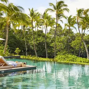 Poolside loungers with umbrellas and palm trees reflected in the water.