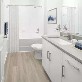 Bathroom with curved shower rod, white cabinets, taupe countertops, and wood-style flooring at Camden Shadow Brook apartments in Austin, TX