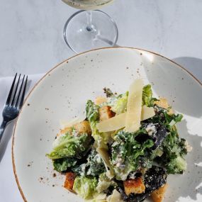 Close up of a Caesar salad topped with shaved parmesan cheese and served with a glass of wine