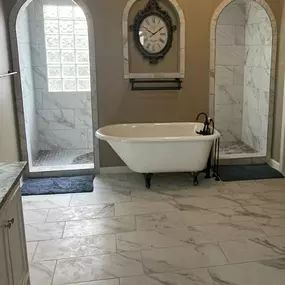 Elegant master bathroom in Overland Park, KS featuring a classic clawfoot soaking tub with dark bronze fixtures, centered between two arched, marble-tiled walk-in shower alcoves. The space is accented by a vintage wall clock framed within a decorative niche, with neutral-toned walls and large-format