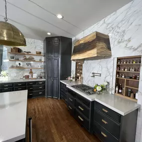 Stunning custom kitchen in Lenexa, KS featuring dramatic full-height marble slab backsplash walls and rich dark cabinetry with brass hardware. A natural wood statement range hood adds warmth and character above a sleek gas cooktop with a built-in pot filler. Open wood shelving displays curated kitch