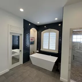 Modern, luxurious bathroom in Lenexa, KS featuring a sleek freestanding soaking tub with matte black fixtures, set against a dramatic navy textured tile accent wall. A large arched window with white plantation shutters adds natural light and elegance. The walk-in shower showcases a herringbone-patte