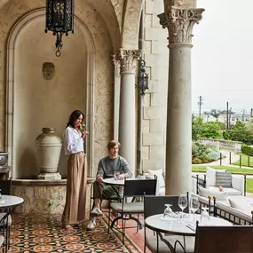 Woman standing on stone terrace with dining tables overlooking a courtyard.