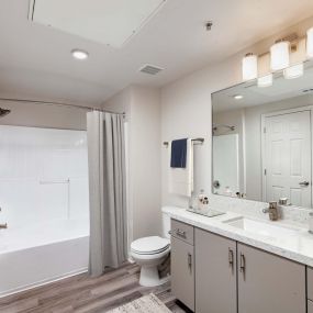Contemporary bathroom with white quartz countertop, greige cabinetry, brushed nickel fixtures, curved shower rod, and wood-style flooring