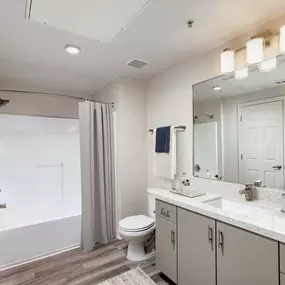 Contemporary bathroom with white quartz countertop, greige cabinetry, brushed nickel fixtures, curved shower rod, and wood-style flooring