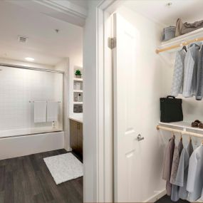 Camden Main and Jamboree Apartments Irvine CA Spacious walk-in closet with wooden shelves and hanger rods near ensuite bathroom with wood-like flooring, white quartz countertops, linen shelves, and glass shower door