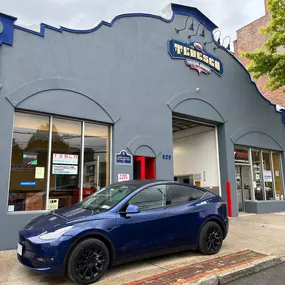 The photo depicts the Tedesco Auto Body building in New Rochelle, NY, with its front entrance standing proudly in full view. The building's façade is painted light grey, and its large glass windows allow a clear view of the showroom inside.