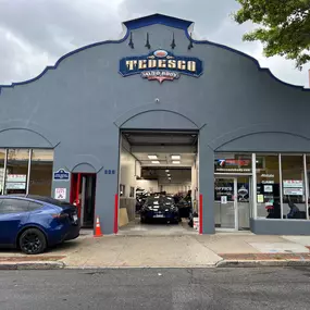 The photo depicts the Tedesco Auto Body building head-on located in New Rochelle, NY, with its front entrance standing proudly in full view. The building's façade is painted light grey, and its large windows allow a clear view of the showroom.