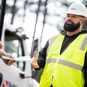 A Builders FirstSource construction worker wearing a white hard hat and neon yellow safety vest receives delivery paperwork outside a jobsite. A branded service truck and trees are visible in the background, emphasizing field communication, customer service, and on-site materials management.