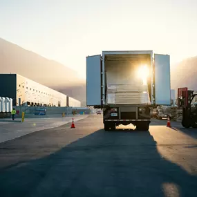 A commercial delivery truck loaded with lumber is parked at a Builders FirstSource distribution center at sunrise, with a forklift actively unloading materials. The image captures the start of a productive day in building materials logistics and jobsite supply chain operations.