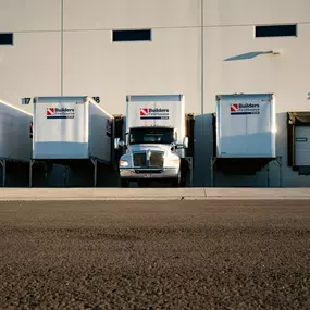 Builders FirstSource semi-truck docked at a warehouse loading bay with multiple trailers lined up in a row.
