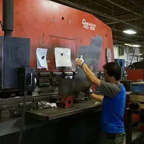 A technician is using an Amada RG-100 press brake machine to bend a large sheet of metal. He is wearing gloves and a blue shirt, standing in front of the industrial forming equipment while carefully aligning the material. The machine’s red housing and multiple forming dies are visible, along with surrounding workstations, pallets, and industrial lighting overhead.