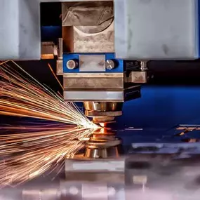 A high-powered laser cutting machine is slicing through a stainless steel sheet, producing a shower of bright orange sparks that illuminate the work area. The laser head is in sharp focus, showing detailed mechanical components and precise alignment. The reflective surface of the metal captures the light from the sparks, while the background remains slightly blurred, emphasizing the cutting action.