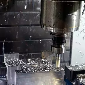 A CNC milling machine is actively machining a block of aluminum, with cutting tools shaping the material. The spindle is engaged in the cutting process, producing bright metallic chips that scatter across the workpiece and surrounding fixture. Coolant spray and fine metal debris coat the machine interior. The background shows the enclosed machining chamber with reflective surfaces from freshly cut aluminum.