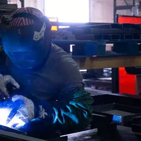 A welder is engaged in MIG welding at a fabrication workstation inside an industrial metal shop. He is wearing a protective welding helmet, safety gloves, and a long-sleeve shirt. A bright blue welding arc illuminates the workspace as he works on steel parts secured to a workbench. The background shows stacked metal beams, pallet racks, and red welding safety curtains, with workshop tools and materials visible.
