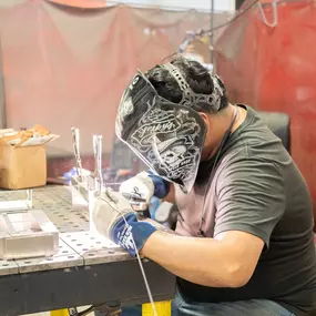 A welder is seated at a metal fabrication workbench, carefully welding aluminum or stainless-steel components. He is wearing a dark t-shirt, protective gloves, and a black custom-designed welding helmet with detailed artwork. The workspace includes a perforated welding table with multiple metal pieces and clamps, as well as a cardboard box and tools in the background. Red welding screens surround the area for safety, and various workshop equipment is visible in the background.