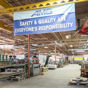 The interior of the AMS metal fabrication facility shows a spacious industrial workspace equipped with heavy-duty machinery, organized sheet metal storage racks, and workstations. A large blue banner reading “Safety & Quality Are Everyone’s Responsibility” hangs prominently from a yellow steel beam. The high-ceiling shop floor features overhead lighting, industrial fans, and palletized materials ready for production. Employees are visible in the background, engaged in fabrication activities.