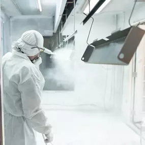 A powder coating technician, dressed in a full white protective suit, hood, gloves, and respirator, applies a fine layer of white powder coating to hanging metal parts inside a controlled spray booth. The metal pieces are suspended from an overhead conveyor, allowing even coverage on all sides. The booth is brightly lit, with powder particles visible in the air, creating a clean, high-contrast industrial scene.