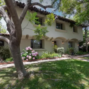 Two-story light stucco building with arched entryway, surrounded by trees, green lawn, and flowers.