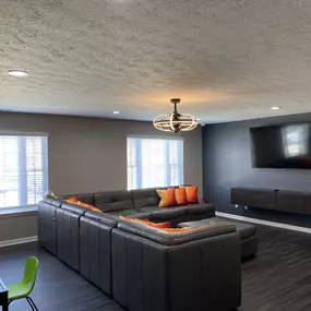 A spacious media lounge at The Park at Greenhill featuring a large sectional sofa with colorful pillows, mounted flat-screen TV, dark wood flooring, modern light fixture, and ample natural light from two large windows.