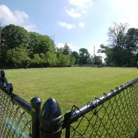 A large, fenced-in bark park at The Park at Greenhill featuring well-maintained grassy space surrounded by mature trees, designed for safe off-leash play and socializing for resident dogs.