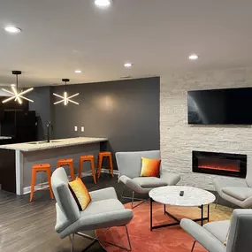 A modern and stylish resident lounge at The Park at Greenhill in West Indianapolis, featuring sleek gray lounge chairs, vibrant accent pillows, a wall-mounted flat-screen TV above a stone fireplace, and a kitchen area with granite countertops, orange barstools, and pendant lighting.