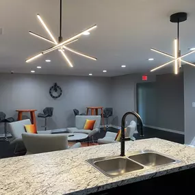 A stylish view of the resident lounge at The Park at Greenhill, featuring modern lighting fixtures, granite countertops, a stainless steel double sink, high-top tables with barstools, and cozy seating with accent pillows in a neutral-toned interior.