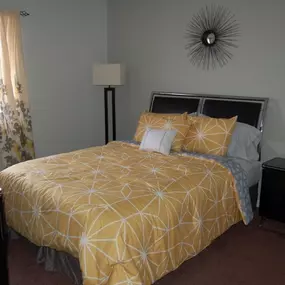 Cozy and well-lit bedroom at The Park at Greenhill, featuring a queen-size bed with geometric bedding, modern black furniture, plush carpeting, and a large window with floral curtains.