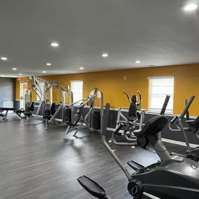 A bright, spacious fitness center at The Park at Greenhill featuring cardio equipment, strength machines, weight benches, and a bold yellow accent wall, with ample natural light and sleek, modern flooring.