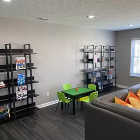 A cozy corner in the community lounge at The Park at Greenhill, featuring open black shelving filled with children’s books and games, a small green play table with matching chairs, and part of a modern sectional sofa with colorful accent pillows.