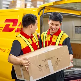 Two DHL Express couriers are handling a large cardboard box during a parcel loading process beside a yellow DHL Express delivery van.
