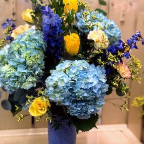 Blue Skies are the best part of a day, so why keep it outside? Sweet blue Hydrangeas accented with golden Roses and deep blue Delphinium. Blue vase may vary.