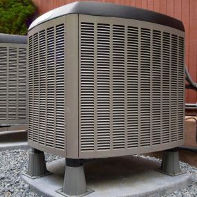 An outdoor air conditioning condenser unit is pictured on a concrete slab surrounded by grass. The unit is a large metal box with vents and a fan on top, and a series of fins on the sides. The fins are coated with a thin layer of dust and debris. The unit is connected to a set of pipes and wires that run along the ground towards the building. The sky above is blue with scattered clouds