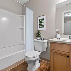 This beautifully renovated bathroom at Water’s Edge Apartments in South Lyon, MI showcases a clean and inviting design, featuring a white shower and tub combination with built-in shelving, a modern vanity with oak cabinetry and brushed nickel fixtures, rich wood-style flooring, and soft neutral walls. The space is thoughtfully accented with natural lighting, minimalist décor, and elegant touches to provide residents with a comfortable and functional retreat.
