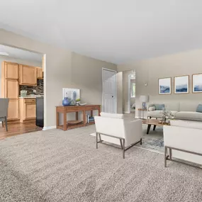 This spacious living room and dining area at Water’s Edge Apartments in South Lyon, MI highlights an open-concept design with plush carpeting, modern neutral wall tones, and a seamless flow into the kitchen featuring warm wood cabinetry and tile backsplash. The room is thoughtfully staged with stylish furniture, elegant lighting, and framed wall art to create a bright and welcoming environment ideal for entertaining or relaxing at home.