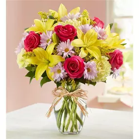 Mom is in for a beautiful surprise! Our best-selling Mother’s Day arrangement is designed in the same tradition as the hand-tied bouquets found in flourishing European flower markets. A vibrant mix of blooms inside a glass vase tied with raffia, it’s a gift that delights with old-world charm and style. All-around arrangement with hot pink roses, yellow Asiatic lilies and carnations and lavender daisy poms; accented with assorted greenery Artistically designed in clear glass vase tied with raffia