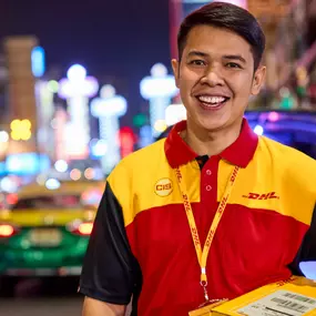 DHL Express courier wearing a branded red and yellow uniform is holding a parcel on a busy city street at night, surrounded by colourful lights and traffic.