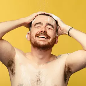 Person with short hair and facial hair smiling and facing the camera. They are washing their hair with a product that creates lather.