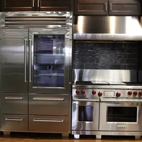 front view of a kitchen display with a stainless steel refrigerator, kitchen range, hood, and oven