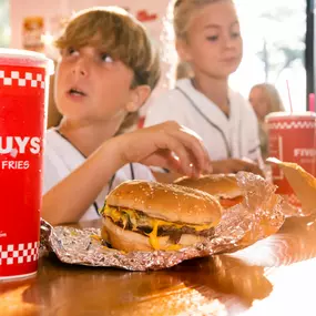 A red Five Guys soft drink cup is the focus in the foreground, as children enjoy their meals in the background.