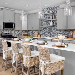Kitchen with gray cabinets, stainless steel appliances with a large kitchen island and 4 woven bar stools wit tile backsplash in DRB Homes Grandview community