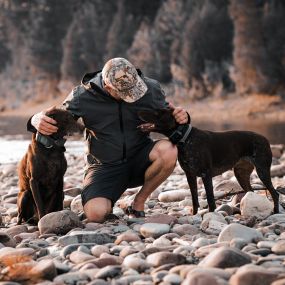 Man crouched down on rocks next to body of water petting two dogs, wearing a FORLOH Airalite Rain Jacket.