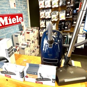 In Imperial Vacuum and Appliances, Bozeman, a Miele canister vacuum is prominently displayed, surrounded by various Miele bags, with the Miele logo against a blue brick wall backdrop.