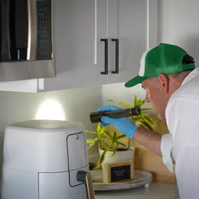 A First Line Pest Defense technician performing a detailed pest inspection under kitchen cabinets with a flashlight.