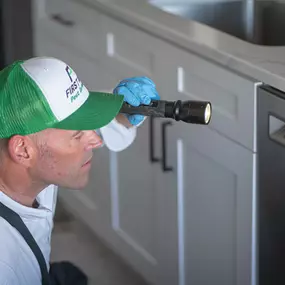 A First Line Pest Defense technician inspecting the area around a kitchen dishwasher with a flashlight for pests.