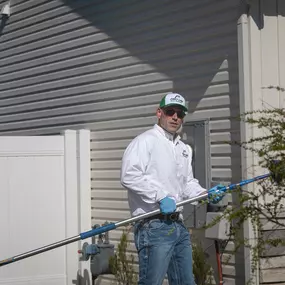 A First Line Pest Defense technician using an extension tool for outdoor pest control on a residential building.